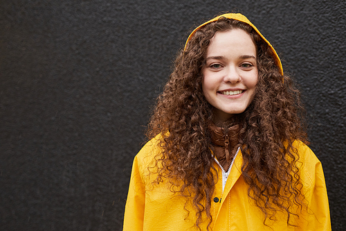 Horizontal medium close-up shot of Caucasian woman wearing yellow raincoat standing against dark gray wall looking at camera