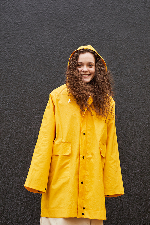 Vertical medium shot of attractive Caucasian woman wearing yellow raincoat standing against dark gray wall looking at camera