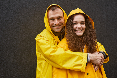 Horizontal portrait of Caucasian man and woman in love wearing yellow raincoats hugging and looking at camera