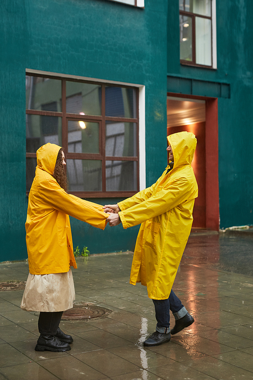 Vertical shot of Caucasian man and woman wearing yellow raincoats having fun holding hands dancing in rain