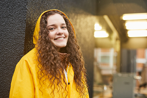Horizontal medium close-up shot of joyful Caucasian woman wearing yellow raincoat standing in rain smiling at camera