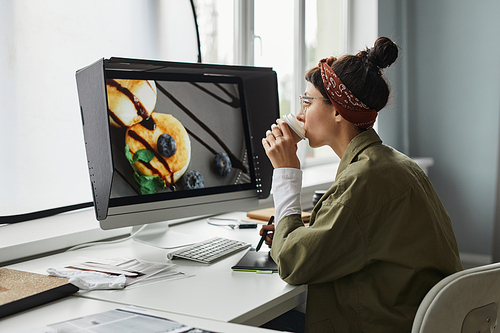 Side view portrait of young female photographer editing photos while using laptop in studio and drinking coffee