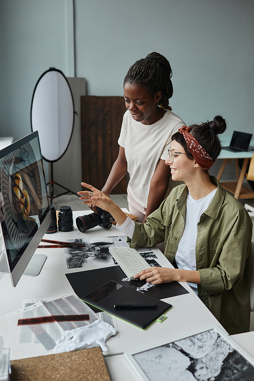 Vertical portrait of two smiling female photographers discussing images on computer screen while working on editing in studio