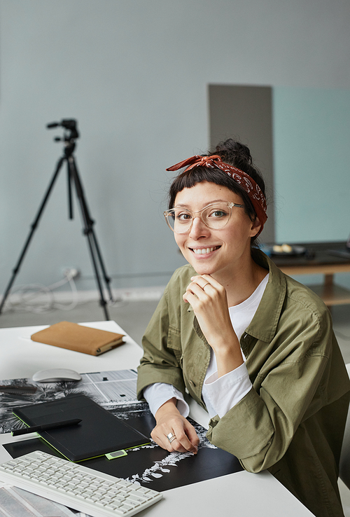 Vertical portrait of young female photographer looking at camera while sitting at workplace in photo studio