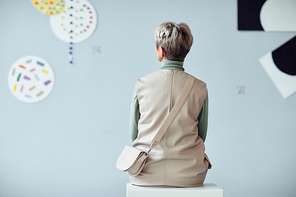 Rear view of unrecognizable mature woman sitting on stool in modern art gallery looking at artworks