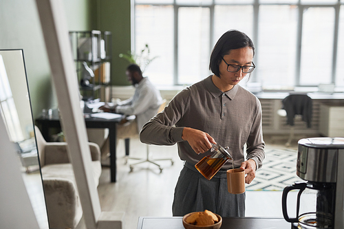 Minimal waist up portrait of young Asian man making coffee in office and wearing grey, copy space