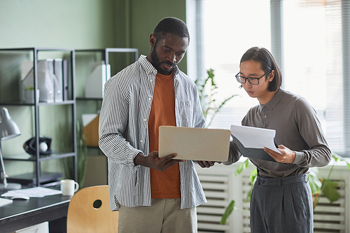 Waist up portrait of two ethnic business people African American and Asian discussing project while standing in modern office