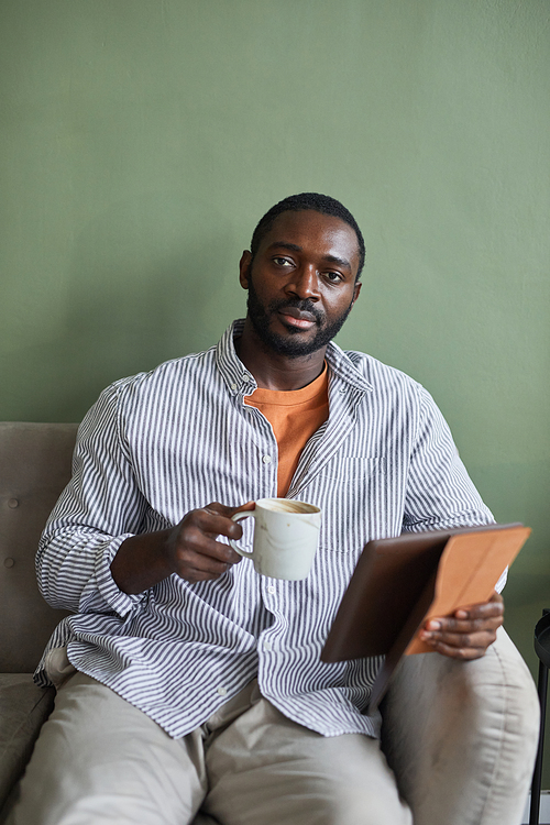 Minimal vertical portrait of handsome African American man holding coffee mug and looking at camera while relaxing on couch against green wall