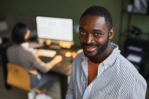 Portrait of modern black businessman smiling at camera while standing in office space with people in background