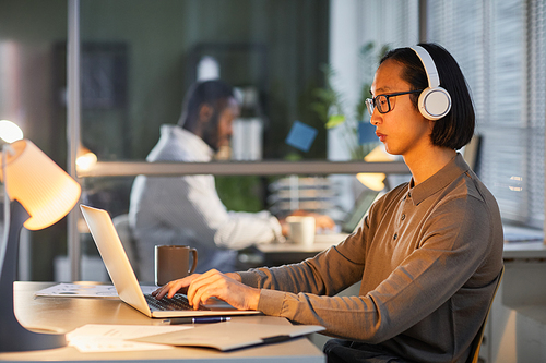 Side view portrait of young Asian man wearing headphones at workplace while working late in office lit by lamps, copy space
