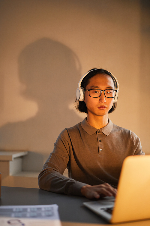 Minimal portrait of young Asian man wearing headphones at workplace while working late in office lit by lamps