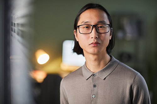 Clean front view portrait of Asian young man looking at camera while standing in office late in evening, copy space