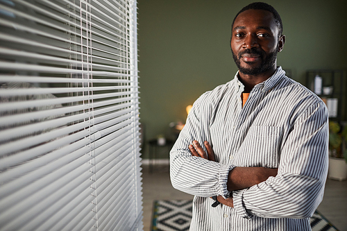 Graphic waist up portrait of successful African American businessman standing with arms crossed in office by window blinds, copy space