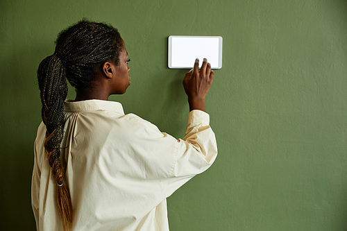 Minimal back view of African American woman using smart home control panel with blank screen on green wall, copy space