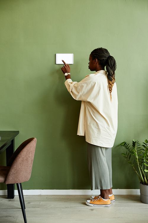 Minimal back view of young African American woman using smart home control panel on green wall