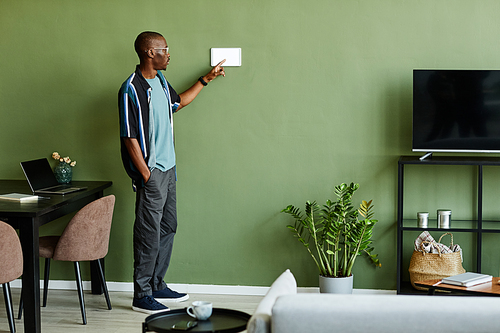 Full length portrait of young African American man using smart home control panel in minimal modern interior, copy space