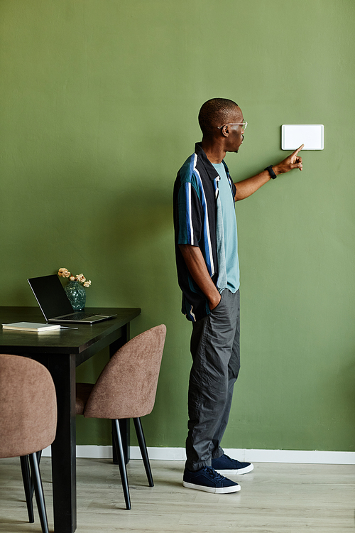 Vertical full length portrait of African American man using smart home control panel in minimal modern interior