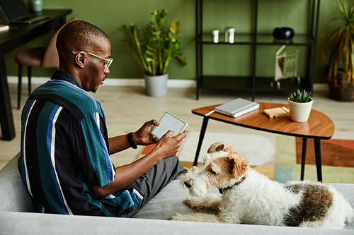 Portrait of black young man holding tablet with blank screen while operating smart home devices in cozy interior with pet dog