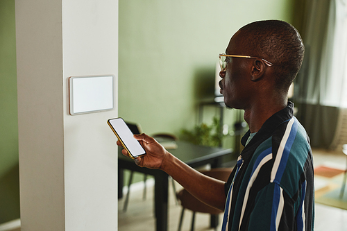 Side view portrait of modern African American man using smartphone while connecting to smart home system