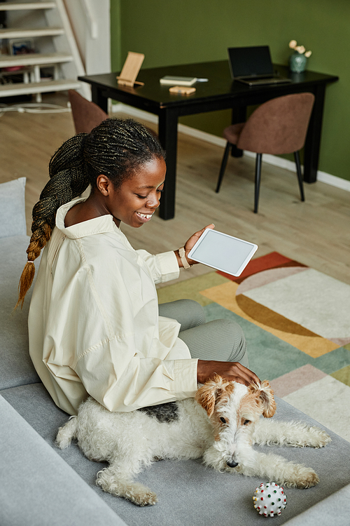 Vertical portrait of smiling black woman petting dog while relaxing on couch at home