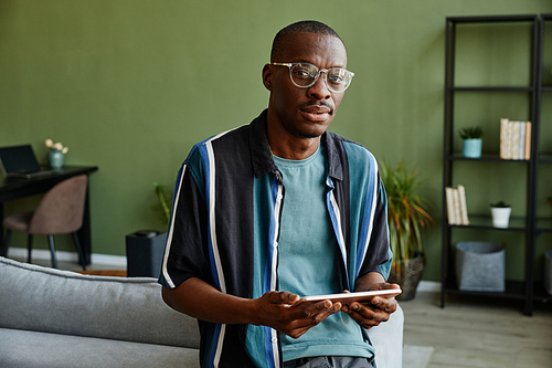 Waist up portrait of modern black man holding tablet and looking at camera in green home interior, copy space