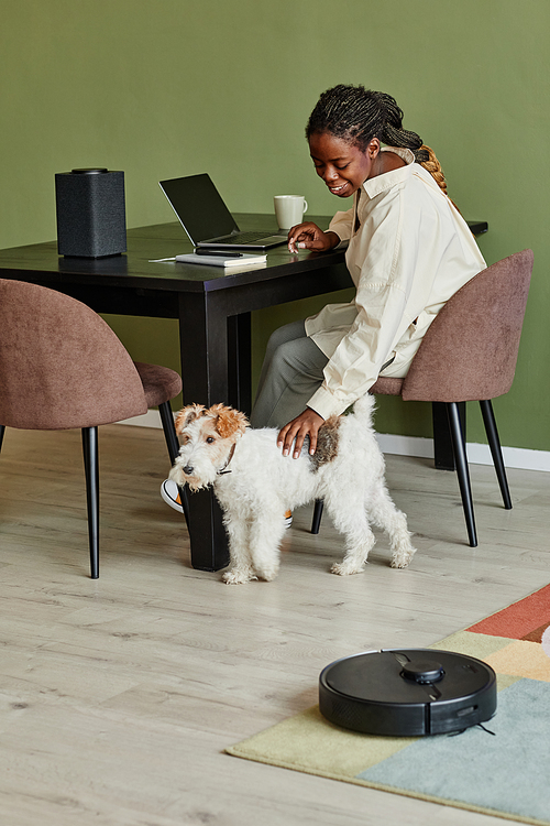 Vertical portrait of young black woman petting dog while working from home in modern apartment