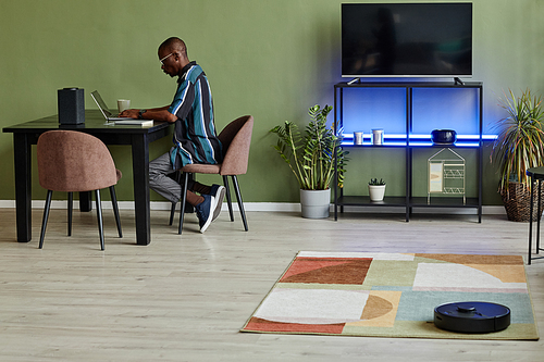 Minimal side view at African American man using laptop at home office with robot vacuum cleaner in foreground, copy space