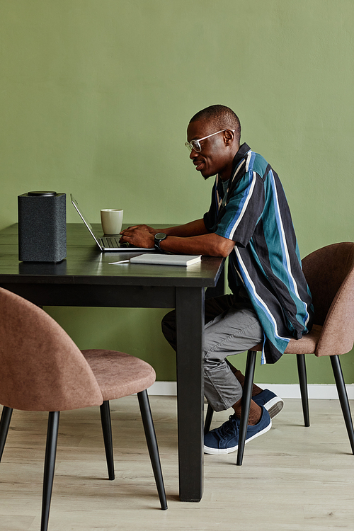Vertical side view portrait African American man using laptop at home office with smart speaker against green wall