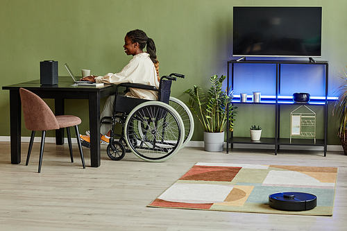 Side view portrait of young African American woman using wheelchair while working at home office with smart home accessible devices