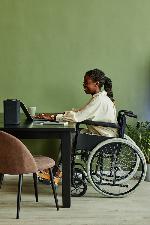 Vertical side view portrait of young African American woman using wheelchair while working at home office against green wall