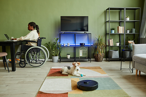 Wide angle view at young African American woman using wheelchair while working at modern home office with smart accessible devices and pet dog