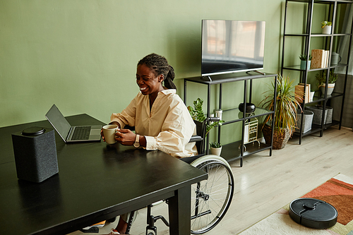 Portrait of smiling of black woman in wheelchair using smart speaker while working from home, copy space