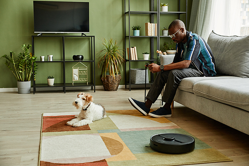 Side view portrait of modern African American man relaxing on couch at home with dog and robot vacuum cleaner in foreground, copy space
