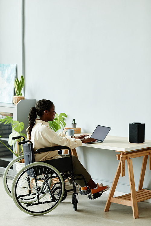 Minimal full length of young black woman in wheelchair using laptop while working at home office, copy space