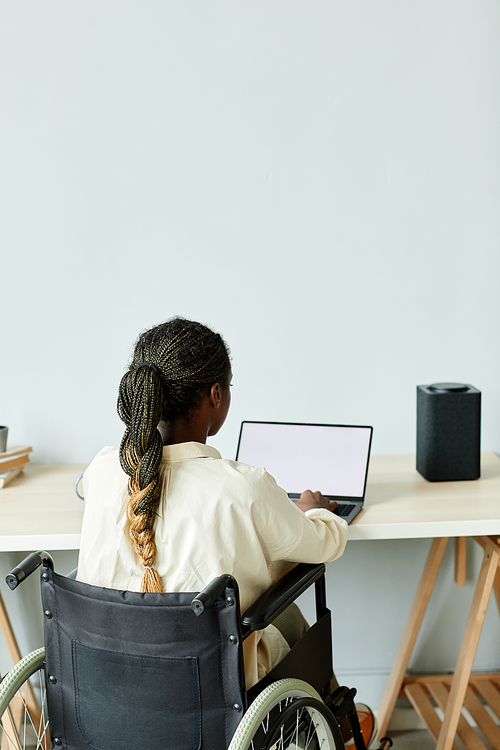 Minimal back view of young African American woman in wheelchair using laptop while working at home office with accessible smart devices