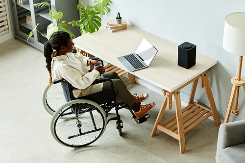 High angle view of young African American woman in wheelchair using laptop while working at home office with accessible smart devices