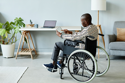 Full length portrait of African American man in wheelchair using tablet with blank screen in minimal home interior, copy space