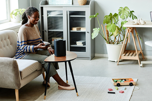 Portrait of smiling African American woman using voice activated smart speaker in cozy home interior, copy space