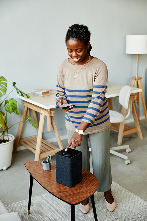 Vertical portrait of smiling African American woman using voice activated smart speaker in modern home