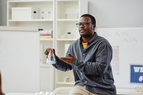 Portrait of African American science teacher explaining chemistry in school class and holding glass beaker with colored liquid, copy space
