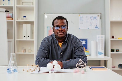 Front view portrait of African American teacher sitting at desk in chemistry class with molecule models, copy space