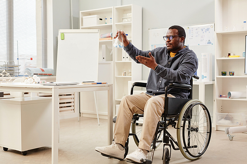 Full length portrait of African American teacher in wheelchair holding glass beaker during chemistry class in school, copy space