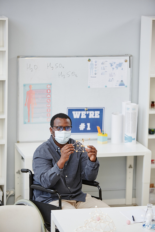 Vertical portrait of African American teacher wearing mask and holding molecule model during chemistry class in school