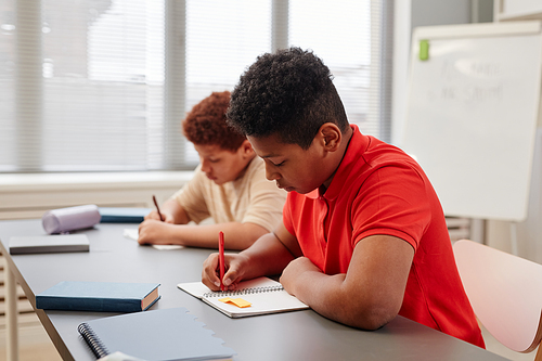Side view portrait of African American teenage boy writing in notebook while studying hard at school, copy space
