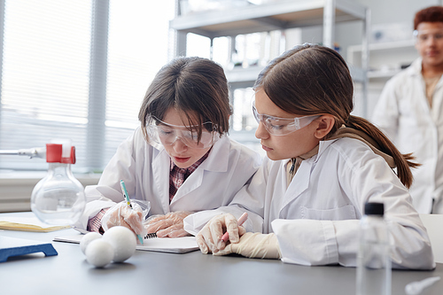 Portrait of two girls taking notes in notebook while doing science experiment in chemistry lab at school