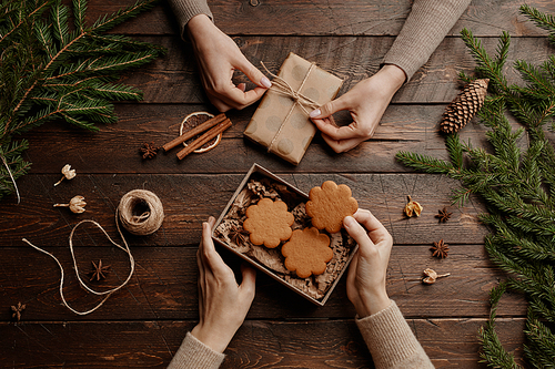 Top view Christmas background with two unrecognizable young women packing homemade cookies in gift wrap at wooden table, copy space
