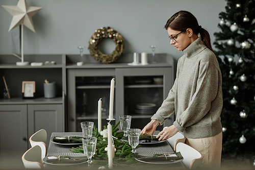 Side view portrait of young woman setting up dining table in dining room decorated for Christmas with fir branches and candles in grey tones, copy space