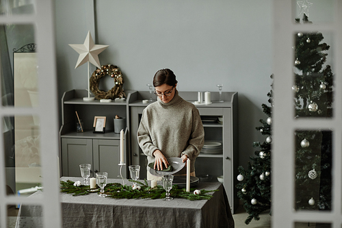 Portrait of young woman setting up dining table in dining room decorated for Christmas with fir branches and candles in grey tones, copy space