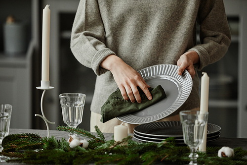 Close up of young woman setting up plates on dining table decorated for Christmas with fir branches and candles in grey tones