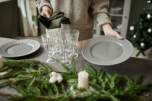 Close up of young woman setting up plates on dining table decorated for Christmas with fir branches and candles in grey tones, copy space